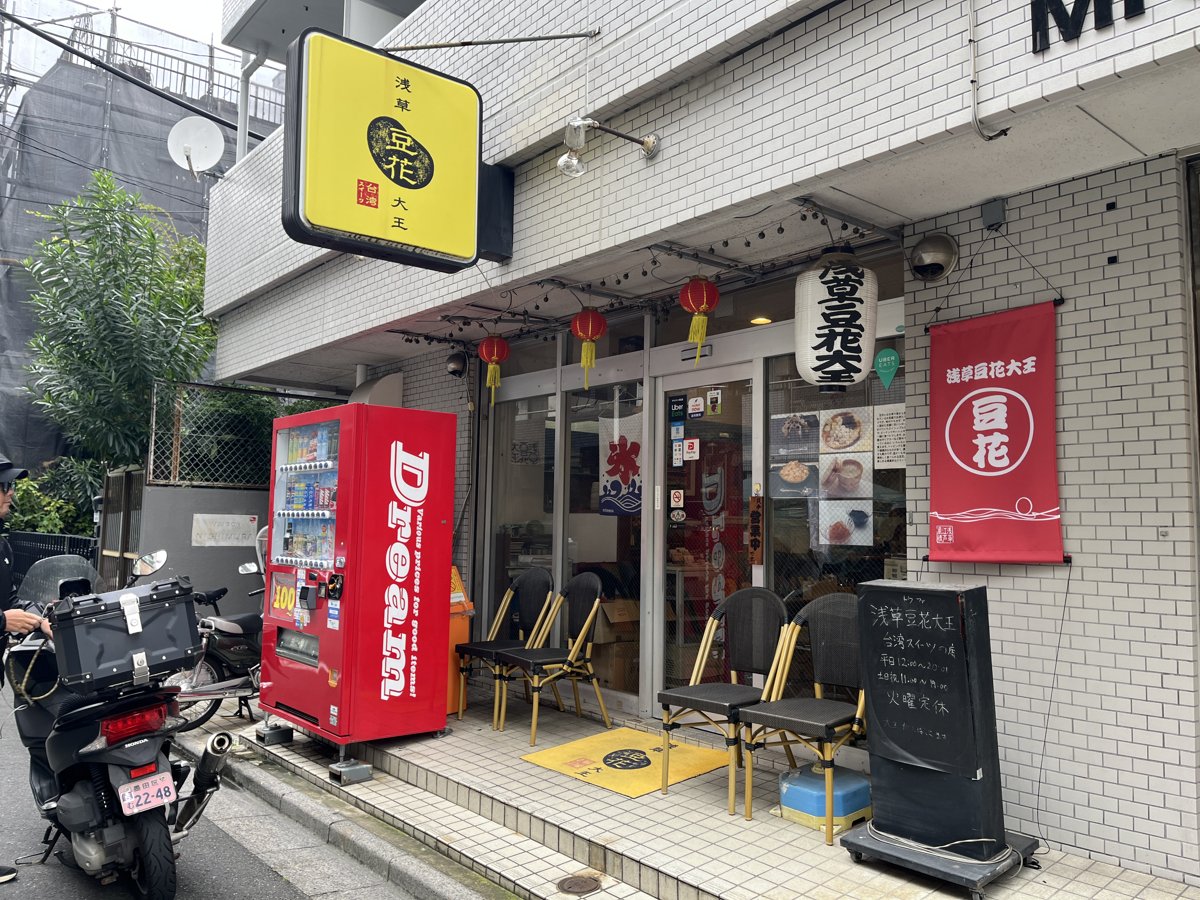 浅草豆花大王の外観。白いタイルの建物の1階に黄色い看板と赤い「豆花」ののぼり、店先に椅子と黒板