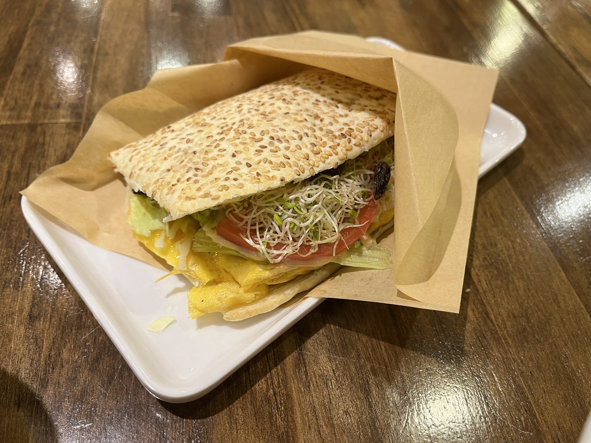 紙に包まれたトマト入り焼餅。ごまのついた平たいパンに、卵焼き、トマト、レタス、スプラウトが挟まっている