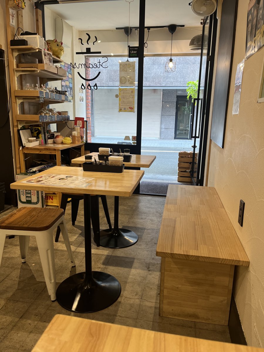 Steamsの店内。木のテーブルと椅子、壁ぎわのベンチ席が並び、窓ぎわに蒸しパンなどを載せた棚とSteamsのロゴがある
